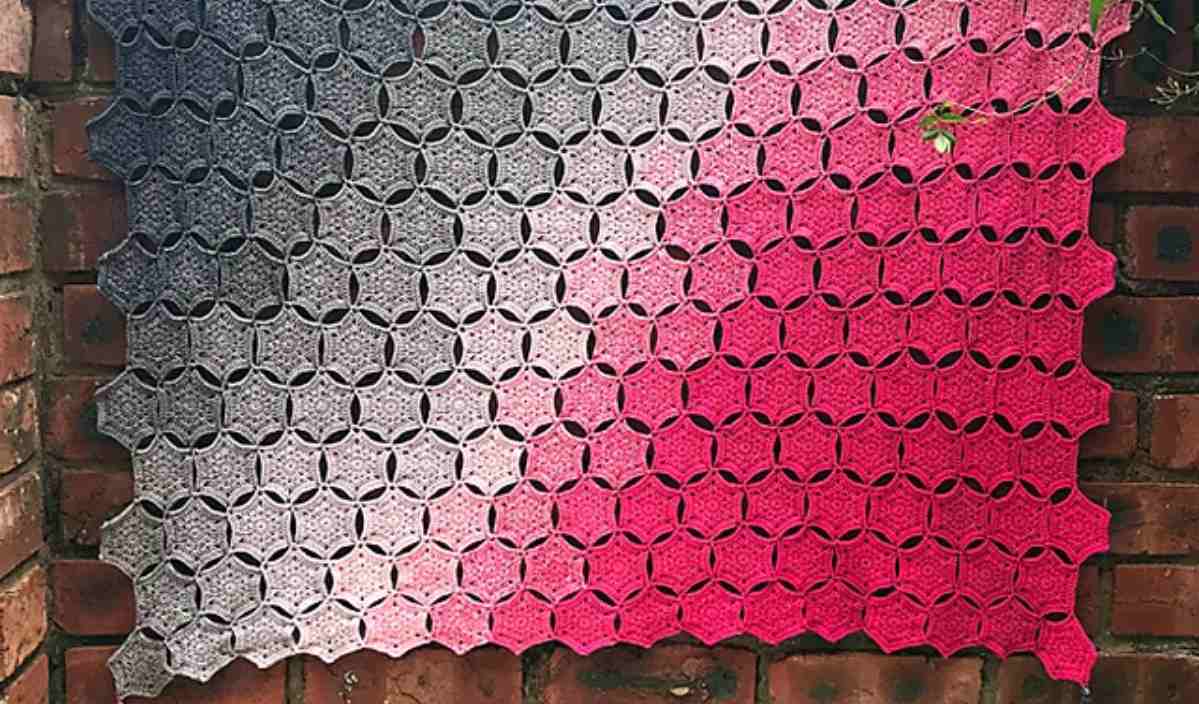 The HexaWhirl Blanket, showcasing a stunning gradient from gray to pink, elegantly hangs on a brick wall. This beautiful crochet piece is inspired by a free crochet pattern that captivates both beginners and seasoned artisans alike.