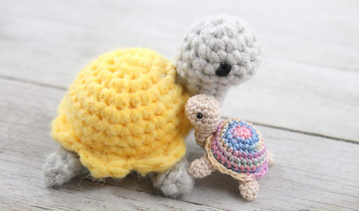 Two crocheted turtles are positioned together on a wooden surface: the larger one in yellow and the smaller sporting a vibrant rainbow blanket shell, perfect for those seeking a free crochet pattern to use up their yarn scraps.