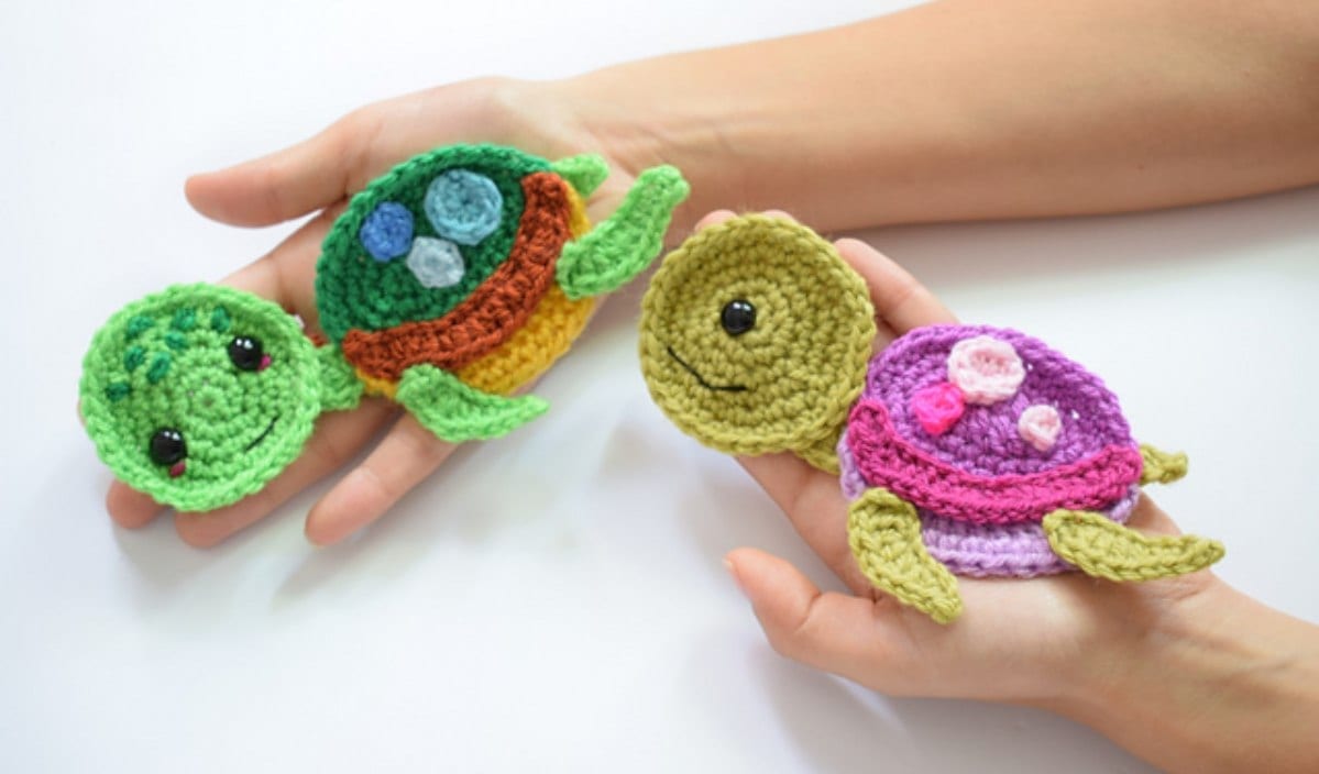 Two hands hold three crocheted turtles in green, brown, and pink, each showcasing unique shell patterns. Perfectly complement your Undersea Sweetness Blanket with these adorable accents inspired by a free crochet pattern.