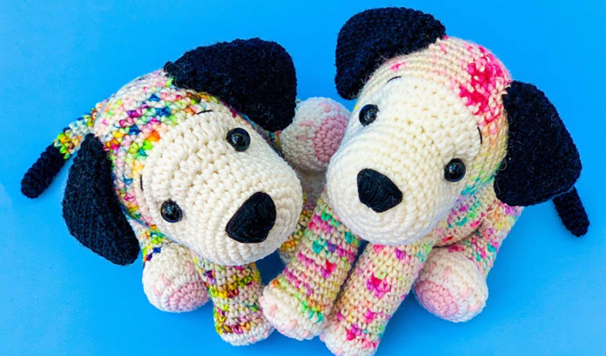 Two crochet dogs with black ears, one multicolored and the other cream with pink spots, are featured against a blue background. This delightful scene is reminiscent of a free crochet pattern design, full of whimsical charm and vibrant creativity.