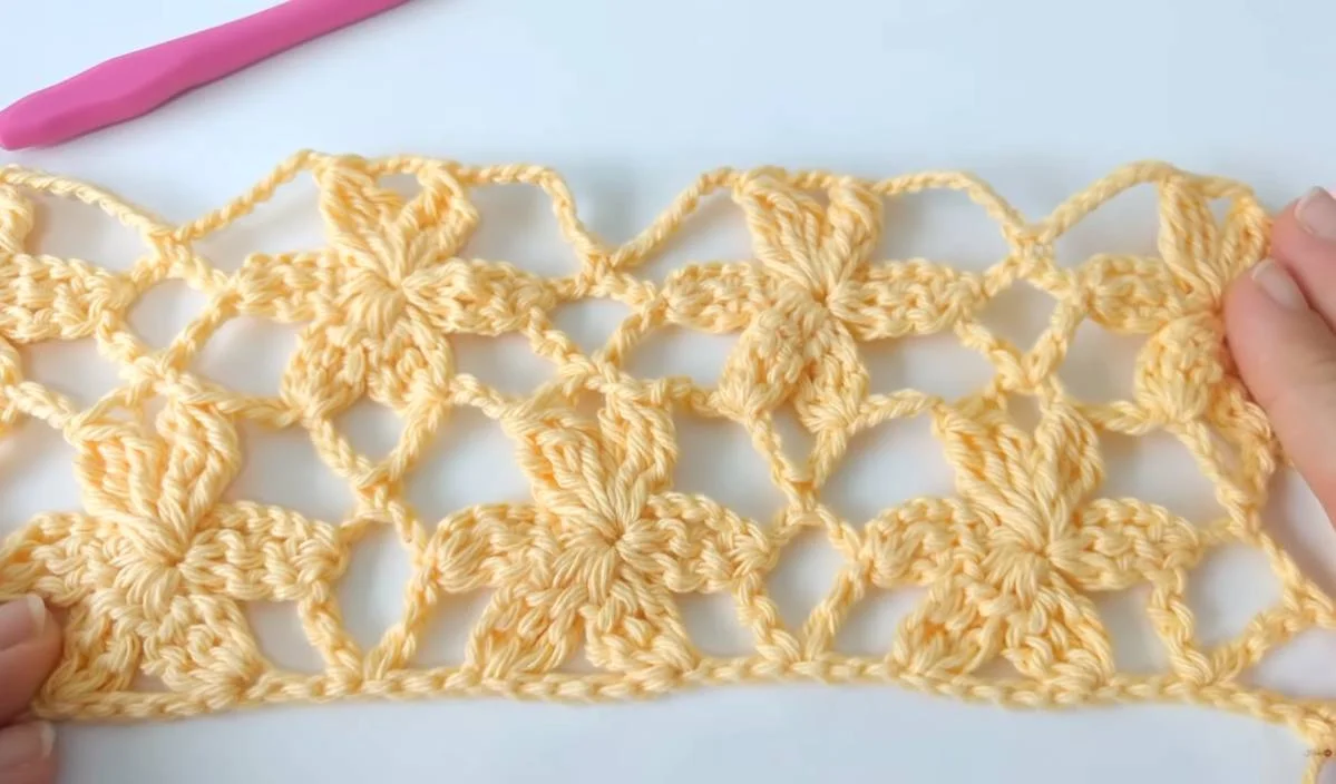 A close-up of a yellow flower mesh crochet pattern with floral motifs, held by hands. A pink crochet hook is visible in the background on a white surface, hinting at this delightful design being part of a free crochet pattern.