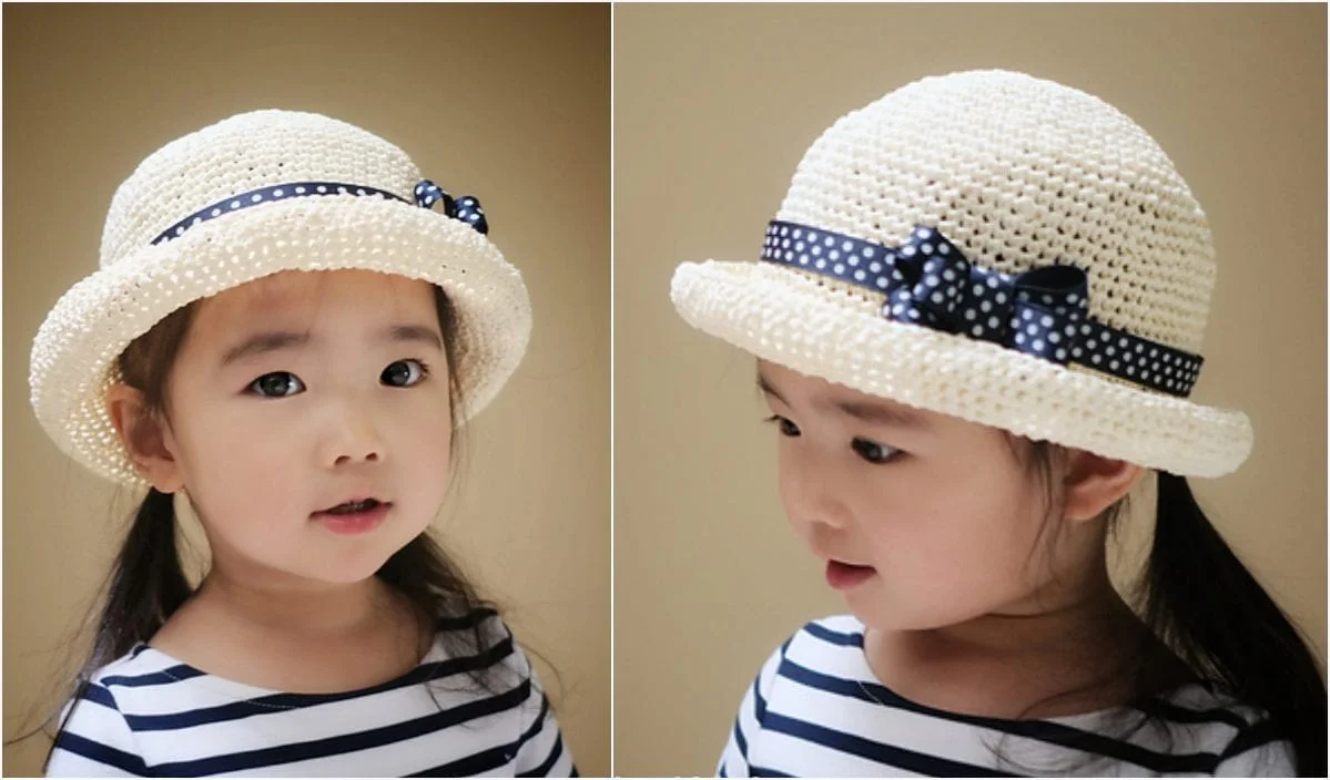 A young child wearing a white woven hat with a black polka-dotted ribbon and adorable Himalayan Rose Slippers glances in different directions. The child is dressed in a striped shirt, completing an endearing look.