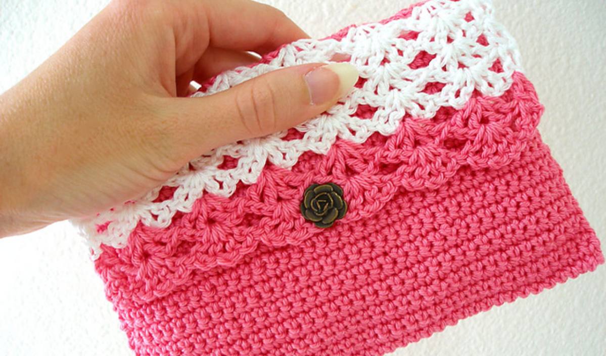 A hand holds a charming pink and white crocheted pouch, adorned with a rose-shaped button, evoking the essence of a delicate flower clutch bag.