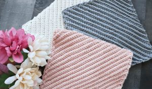 Three knitted washcloths in white, grey, and pink are arranged in a fan shape beside a cluster of pink and white flowers on a dark surface. Find inspiration for your own creations with our free crochet pattern and accompanying video tutorial.