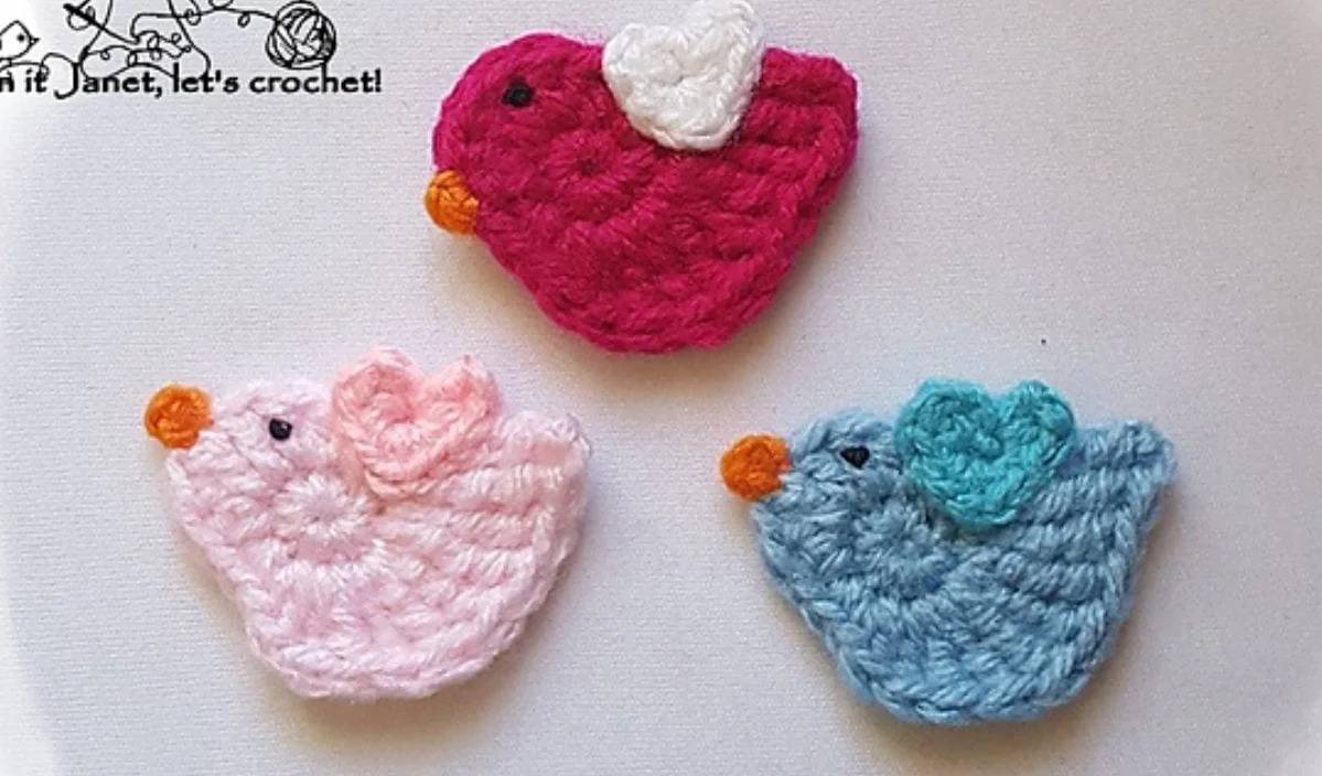 Three vibrant bird appliques, crafted with free crochet patterns in pink, blue, and fuchsia, showcase orange beaks and delicate white, pink, and blue wings. They are artfully arranged on a pristine white surface.