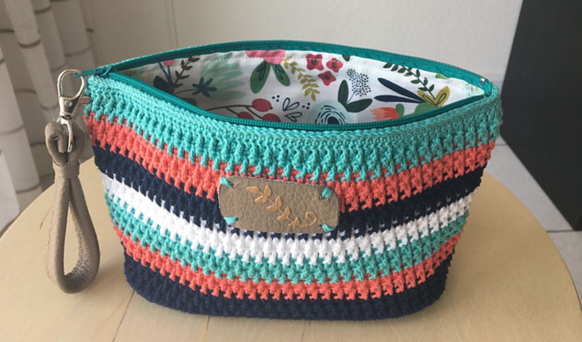 Discover the charm of a crocheted zipper pouch with floral lining, boasting horizontal stripes in teal, coral, navy, and white. Complete with a leather strap on the side, this stylish accessory is perfect for any occasion. Check out our free crochet pattern to create your own!