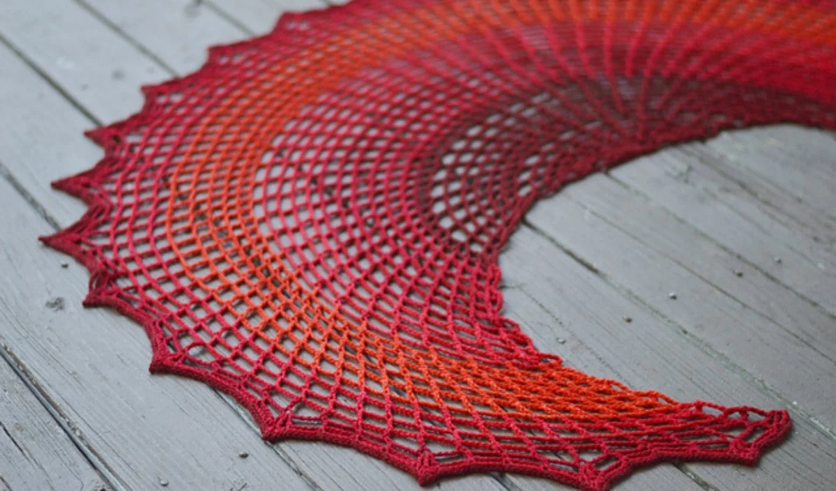 A knitted lace shawl in gradient shades from orange to red, reminiscent of Philip the Dragon's fiery hue, displayed on a gray wooden surface.