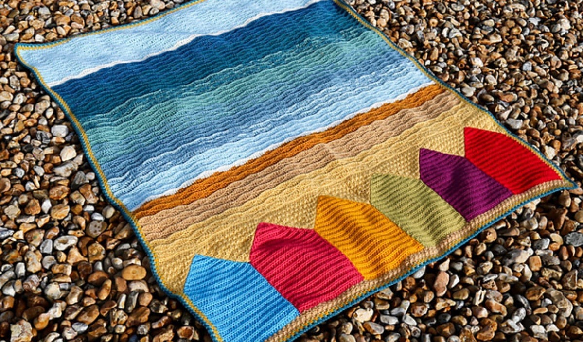 Discover the Swanage Blanket, a crocheted masterpiece capturing a beach landscape on a pebble surface, featuring colorful beach huts and waves. Perfect for summer vibes, this design offers a stunning free crochet pattern that brings coastal charm to any space.