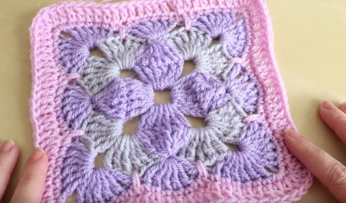 A crochet granny square with a tricolor crochet design, featuring a gray and purple pattern in the center, framed by a pink border. Two hands are holding the edges.