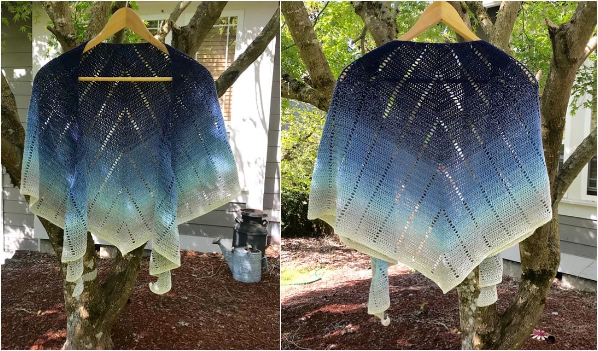 A triangular, knitted croissant shawl with a gradient pattern from navy blue to white, displayed on a hanger outside among tree branches.