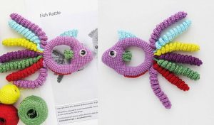Vibrant Fish Baby Rattle featuring a ring body and multicolored spiral fins, beautifully displayed on a white surface next to a free crochet pattern sheet and yarn balls.