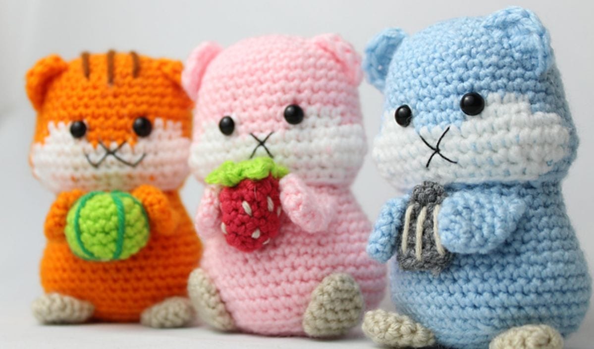 Three crocheted animal figures: an orange one with a green ball, a pink one holding a strawberry, and a blue one with a gray ball. It's snack time in this delightful free crochet pattern featuring charming creations perfect for hamster lovers and crafty enthusiasts alike.