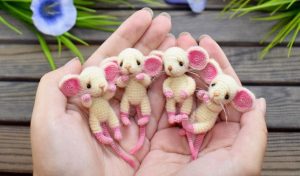 Hands holding four cute amigurumi mice, each with beige bodies, pink ears and feet, on a wooden surface adorned with purple flowers and green stems nearby.