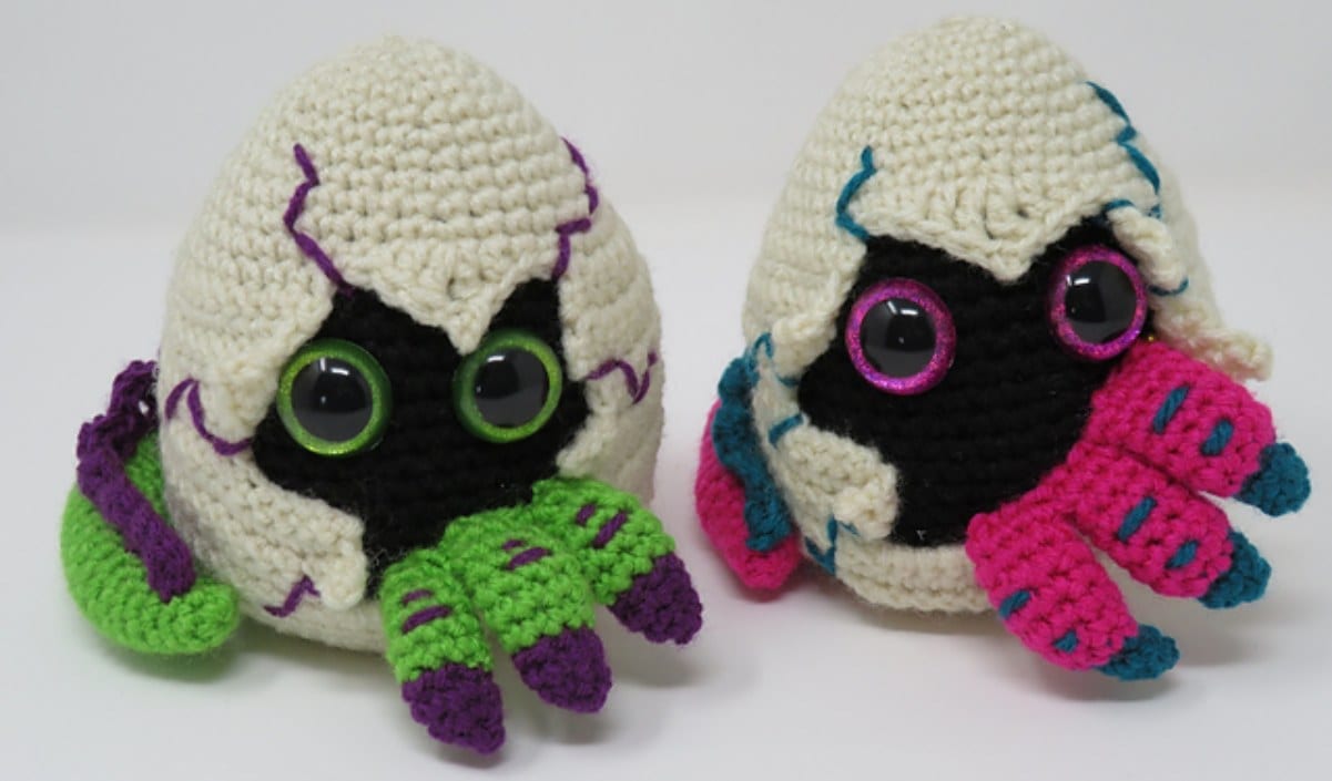 Two crocheted creatures partially emerging from cracked dragon egg shells, featuring large eye patterns and colorful tentacle-like appendages.