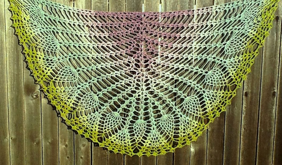 A colorful, intricately crocheted shawl with a gradient pattern, inspired by the Pineapples Shawl design, hangs elegantly on a wooden fence.