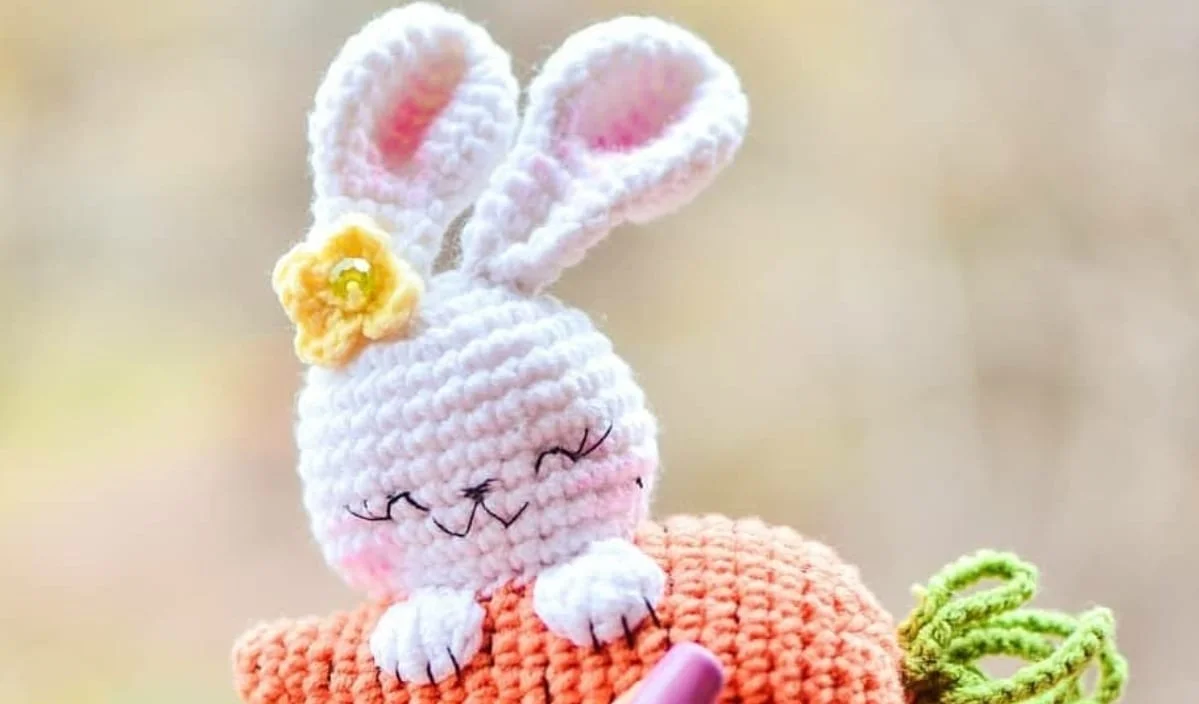 This delightful crocheted white bunny, with its serene closed eyes and a jaunty yellow flower atop its head, holds a large crocheted carrot. It's an adorable creation perfect for those seeking a charming rabbit brooch or eager to try their hand at a free crochet pattern.