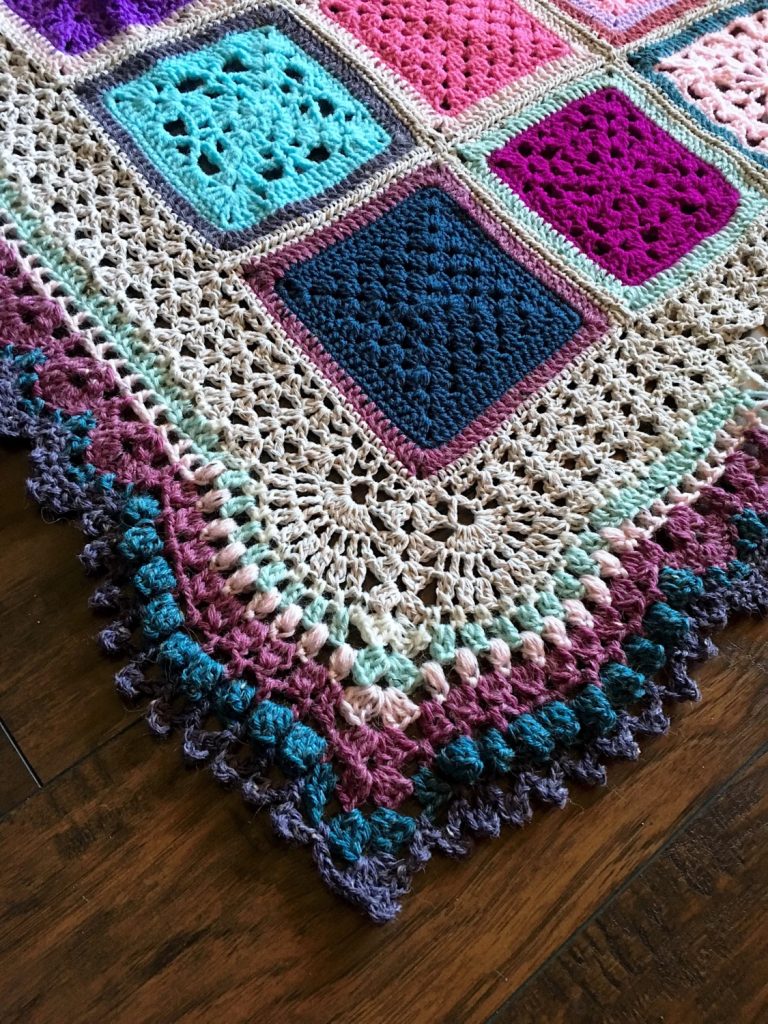A multicolored crocheted blanket featuring square patterns and a detailed crochet blanket border with scalloped edges, displayed on a wooden floor.