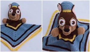 A crocheted Chase Lovey blanket showcases an animal head with large eyes, inspired by the beloved Paw Patrol character. The body features a charming crochet pattern in blue, yellow, brown, and gray square designs.