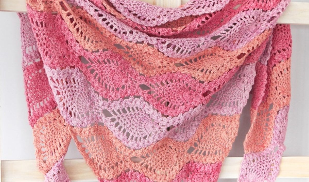 A crocheted shawl with wavy pink, peach, and coral patterns hangs gracefully over a wooden rack, echoing the gentle charm of a rose bunny.