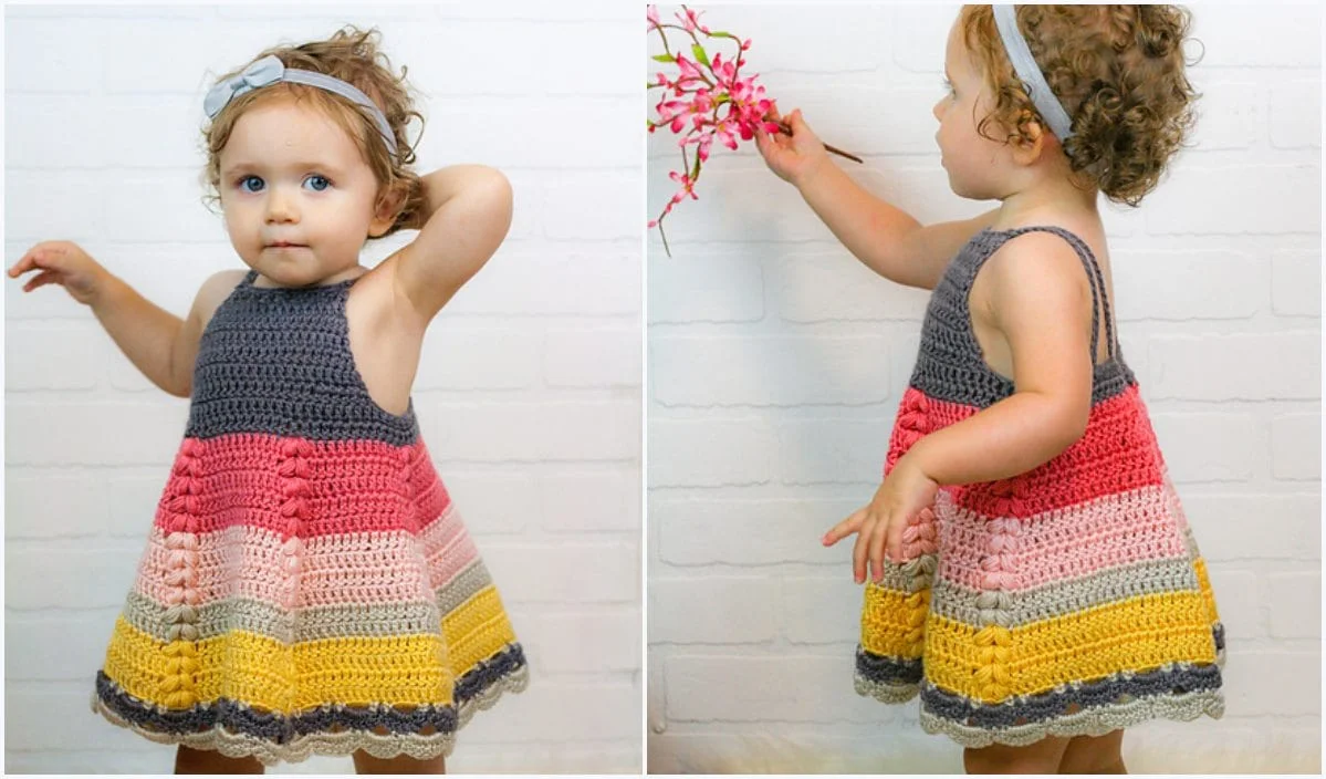 A toddler in a vibrant, puff stitch crocheted dress leans against a white brick wall, clutching a pink flower branch.