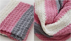 A crocheted blanket with pink, white, and gray stripes is elegantly folded and draped, resembling the cozy charm of a timeless triangle shawl.