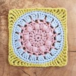 Discover the Calm Cove Square, a crochet granny piece adorned with pink, blue, and green concentric patterns set against a wooden background. Dive into creativity with our free tutorial, perfect for bringing this serene design to life.