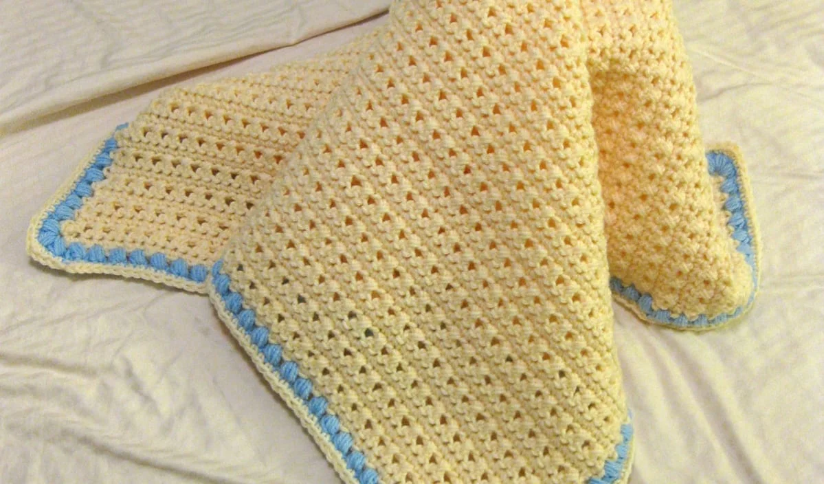 A handmade baby crochet blanket in cream with a blue border is gracefully displayed on a light-colored surface.