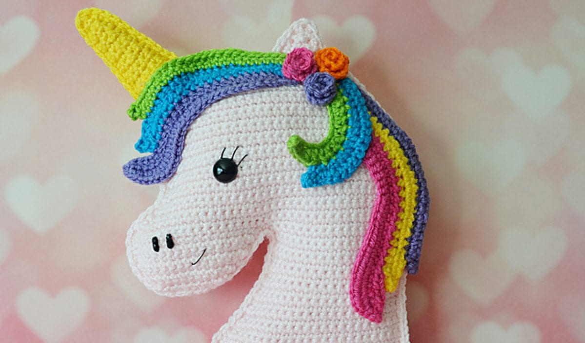 This Kawaii crocheted unicorn head features a yellow horn, rainbow mane, and floral detail, set against a pink heart-patterned background. Perfect for adding a whimsical touch to your space or to inspire your next crochet pattern adventure!