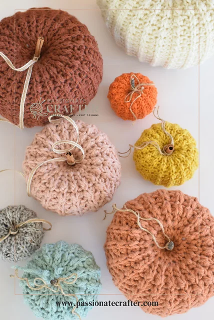 Several crocheted pumpkins of different sizes and colors, each featuring a stick stem and string, are arranged on a white surface. Perfect for autumn dรฉcor, these charming creations showcase the versatility of a crochet pumpkin pattern. Text reads: www.passionatecrafter.com.