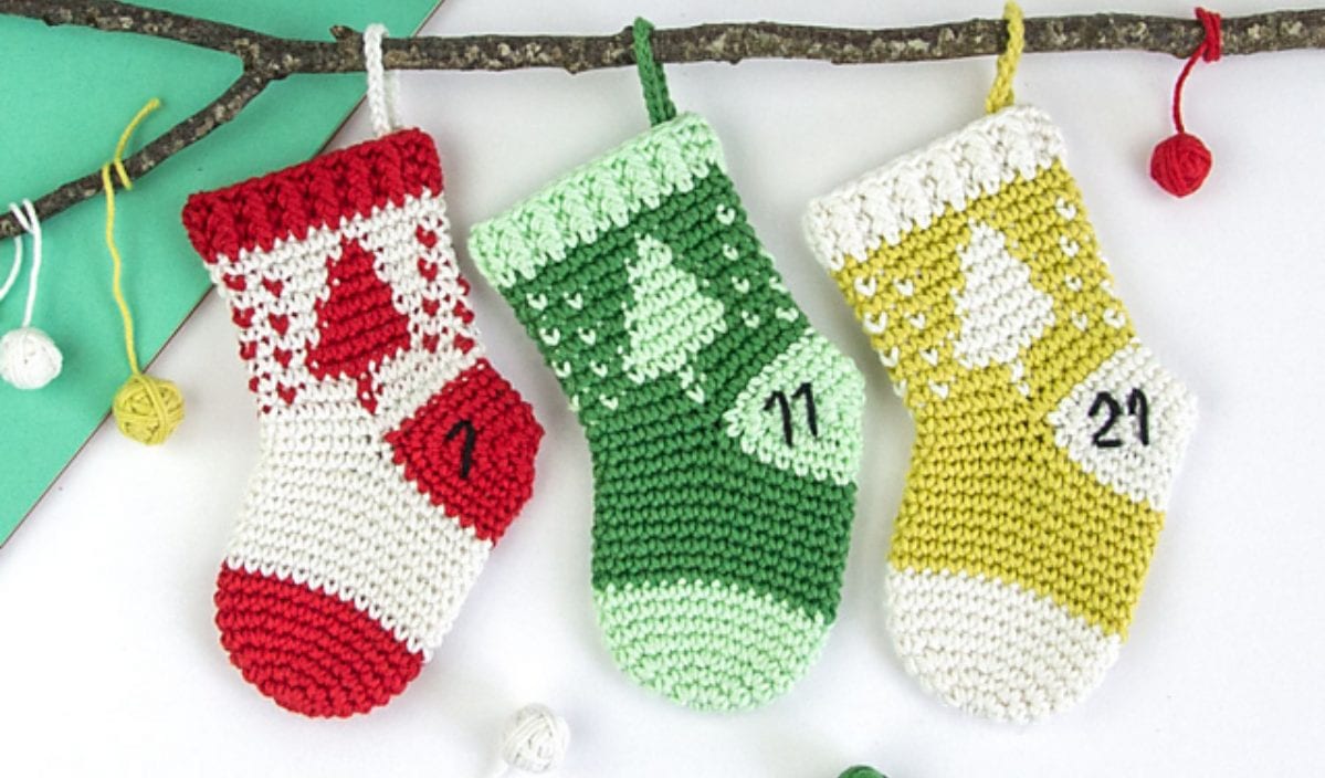 Three crocheted Christmas stockings in vibrant red, green, and yellow hang from a branch. Each stocking features a tree design and is numbered 7, 11, and 21. Perfect for the holiday season, these Christmas stockings can be crafted using free crochet patterns available online.