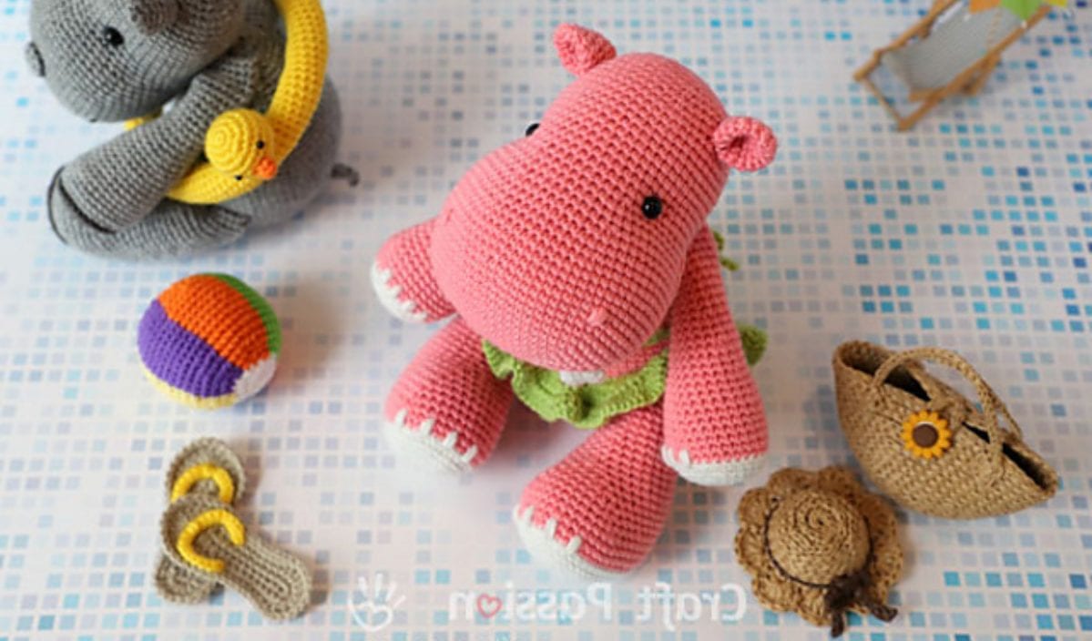 The amigurumi pink hippo toy, donned in green shorts, sits on a patterned surface. It's surrounded by a delightful crocheted elephant, ball, sandals, plaid bag, and hat. Perfect for those seeking inspiration from free crochet patterns!