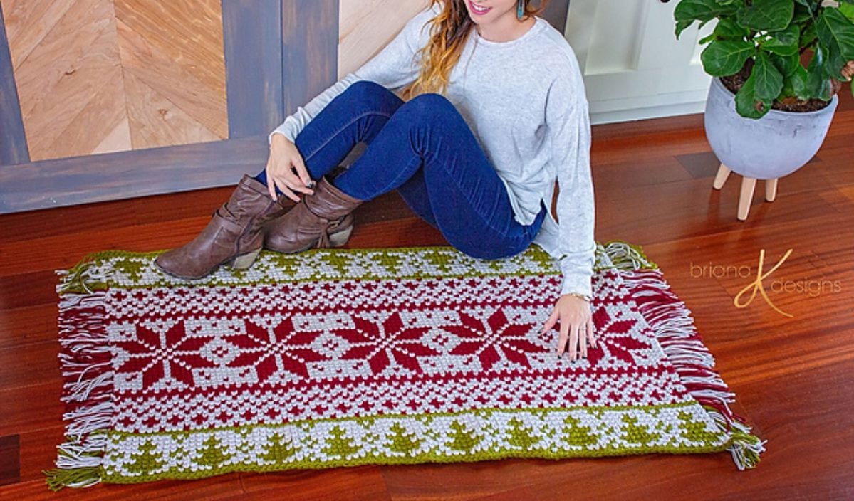 Seated on the floor next to a vibrant green, red, and white patterned rug with fringe edges, the person exudes a serene holiday spirit. A potted plant adds to the cozy scene.