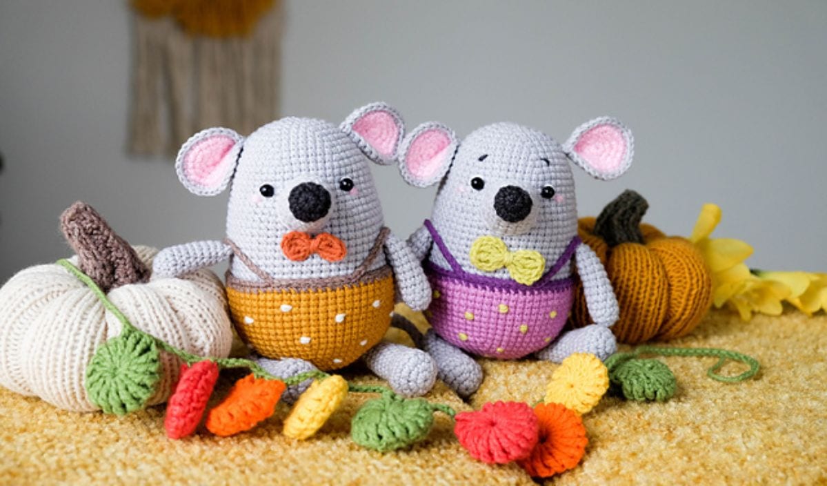 Two crocheted mice with bow ties, made using free crochet patterns, sit among yarn pumpkins and a colorful garland on a yellow surface.