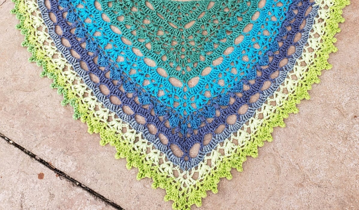 A colorful crocheted triangular shawl, crafted from free crochet patterns, boasts intricate designs in shades of green, blue, and purple. It elegantly rests on a light stone surface.