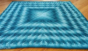 A crocheted blanket featuring an optical illusion effect with a blue gradient pattern is arranged in a symmetrical, geometric design on a wooden floor.
