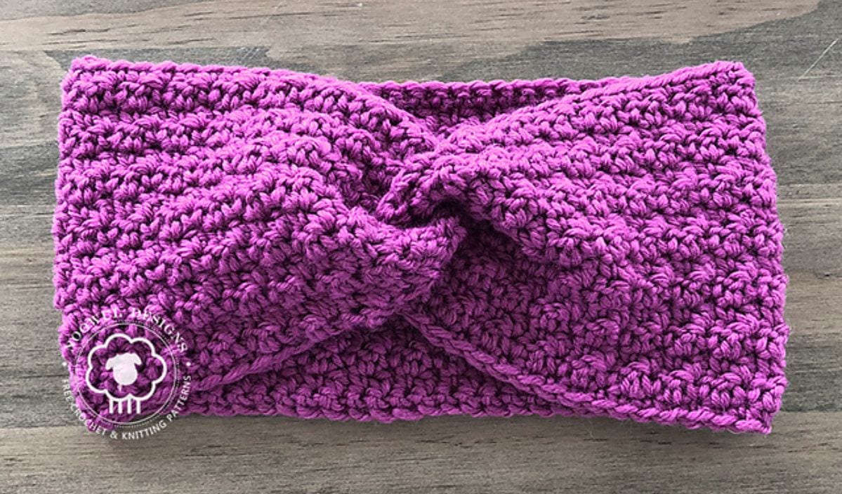 A knitted purple earwarmer featuring a twisted knot design is laid flat on a wooden surface, showcasing its intricate crochet patterns.