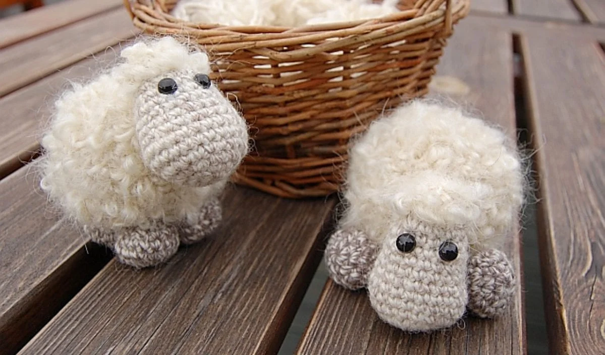 Two crocheted sheep keychain figurines with black eyes sit on a wooden table in front of a wicker basket filled with similar sheep, all crafted from delightful crochet patterns.