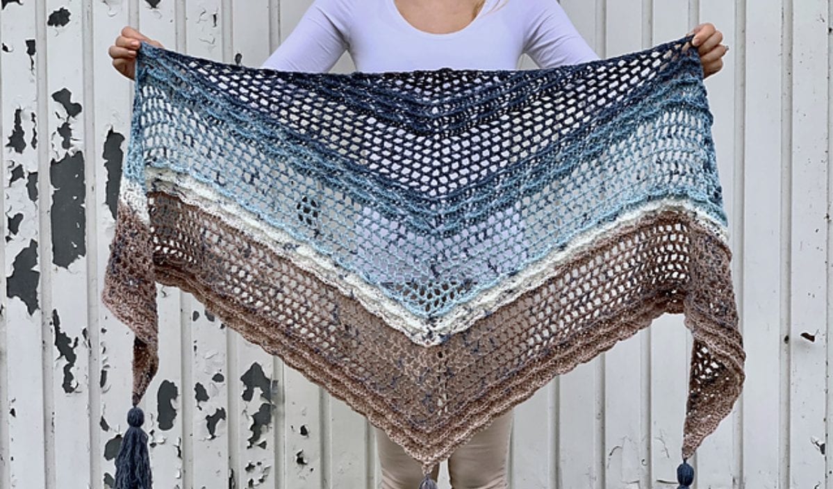 Person holding a handwoven blue and brown triangular Shell Motif Shawl with tassels against a white, weathered wall.