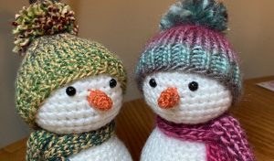 Two tiny crocheted snowmen with orange noses, wearing colorful knit hats and scarves, sit side by side on a wooden surface. Perfect examples of charming crochet patterns for winter decor.