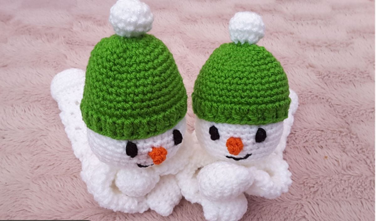 Two crocheted snowmen, crafted from free crochet patterns, with green hats and orange noses lie on a textured pink surface.