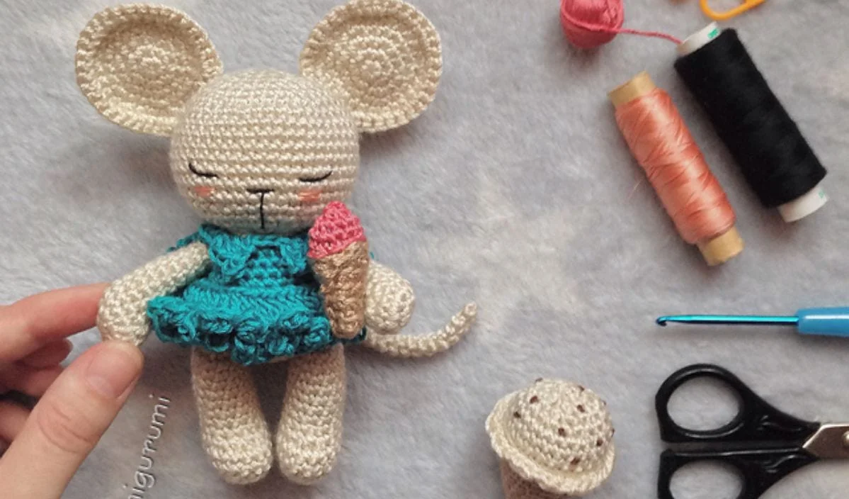 This Sleepy Mouse, dressed in a blue dress and holding an ice cream cone, is nestled among crochet materials—yarn, crochet hook, and scissors—on a gray background. Perfect for those seeking inspiration from adorable crochet patterns.