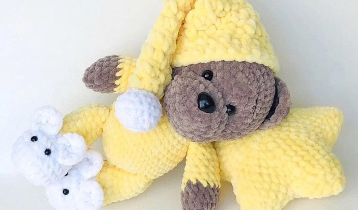 Goodnight Bear is a plush bear clad in yellow pajamas and a hat, clutching a yellow star, and donning white slippers shaped like bear faces. Ideal for those who cherish crochet patterns in their life.