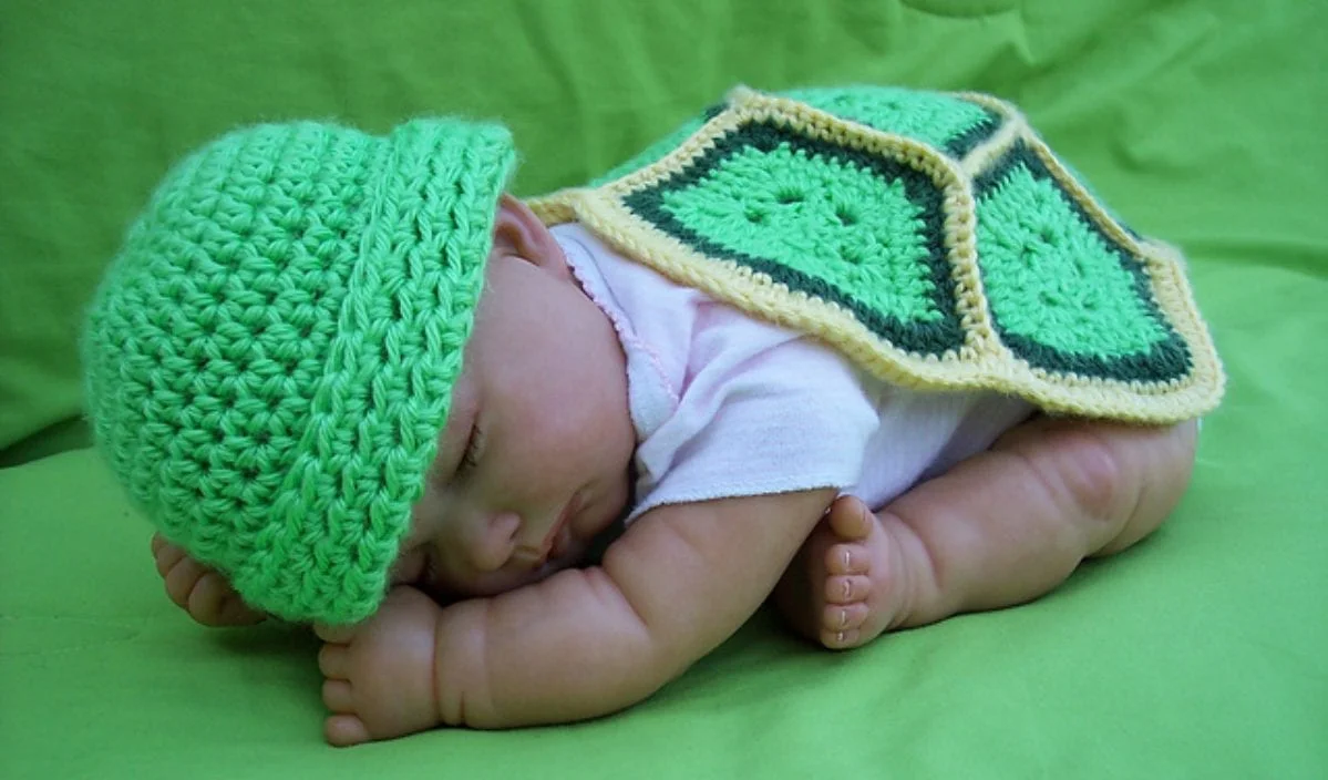 A newborn is peacefully sleeping on a green surface, adorned in a crochet hat and a crocheted turtle shell outfit. This charming photo set highlights intricate crochet patterns that add an adorable touch to the scene.