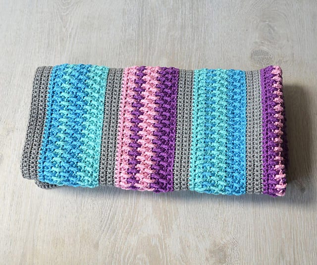 A knitted blanket with horizontal stripes in shades of blue, pink, purple, and gray lies gracefully on a light wooden surface, reminiscent of a free crochet pattern that inspires cozy home decor.