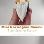 A hand holds a crocheted Norwegian gnome, crafted from a delightful crochet pattern, featuring a yellow hat, white beard, and blue body. Below it, four similar gnomes in various colors are charmingly lined up.