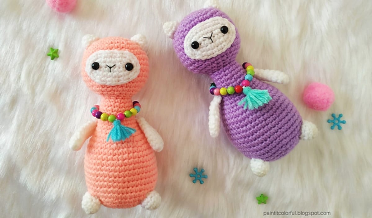 Two crocheted amigurumi llama dolls, one pink and one purple, adorned with bead necklaces and tassels, rest on a white fluffy surface. Colorful decorations surround them, showcasing intricate crochet patterns that bring whimsy and charm to the scene.