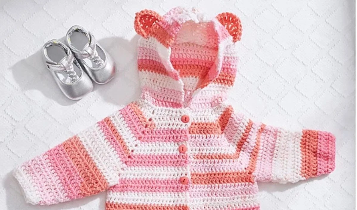 Crocheted pink and white baby jacket with bear ears, reminiscent of free crochet patterns, laid out beside a pair of silver baby shoes on a white textured background. Perfect for those end-of-summer evenings.