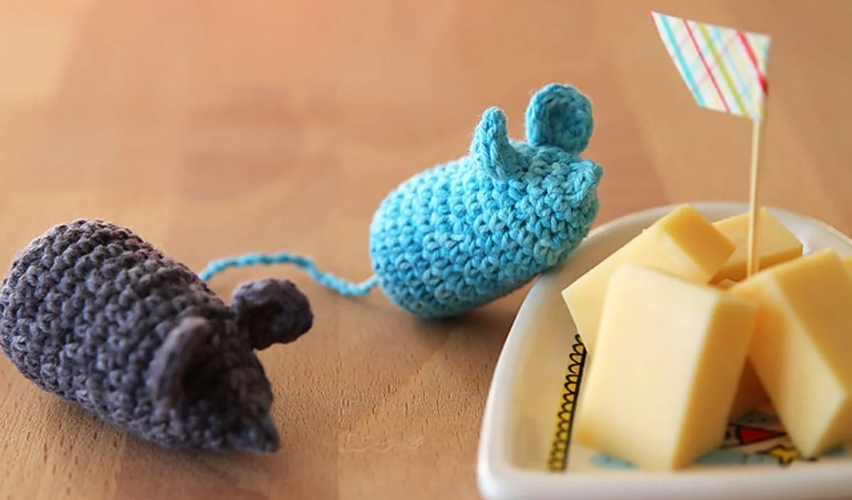 Two knitted mouse toys sit beside a small dish with cubes of cheese and a decorative toothpick, showcasing delightful examples of creative crochet.