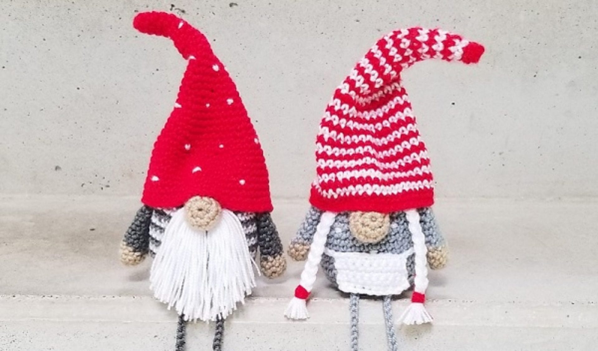 Two knitted gnome figures with long hats and white beards sit on a concrete surface. One sports a vibrant red hat, while the other dons a whimsical red and white striped hat, offering inspiration for gnome-free crochet patterns.