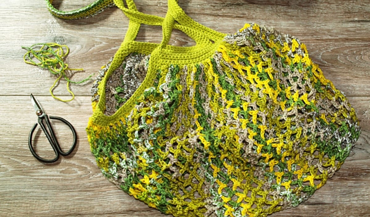 A colorful, crocheted green and yellow mesh bag sits on a wooden surface, accompanied by scissors and yarn, hinting at the creative journey of exploring new crochet patterns.